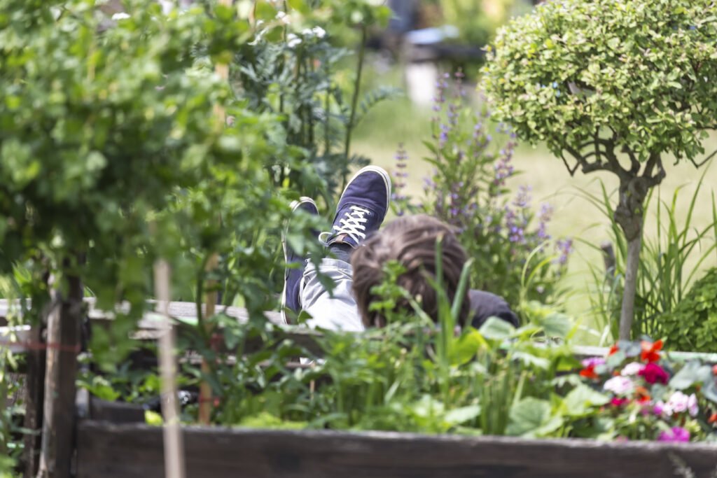 Naure support for burnout: A person resting in a garden with their feet up on a wooden planter, surrounded by lush green plants and purple flowers. A peaceful example of gentle ways to recover from burnout.